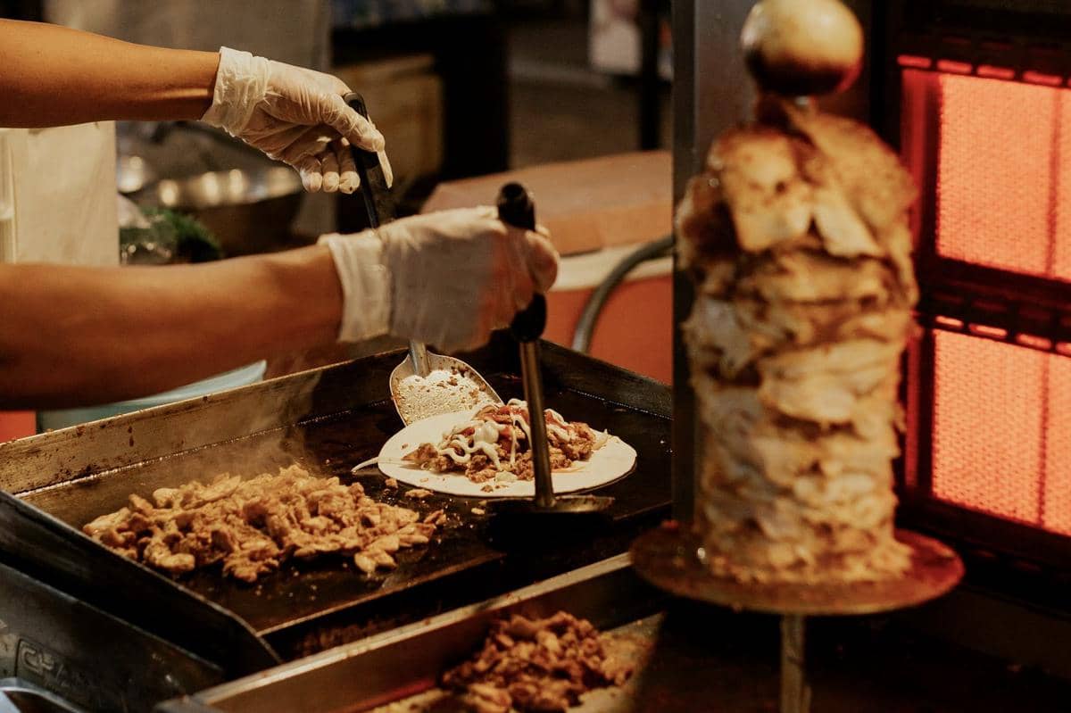 Penjual makanan jalanan sedang menyiapkan daging panggang di atas pemanggang dan menyajikannya di atas roti dengan saus.
