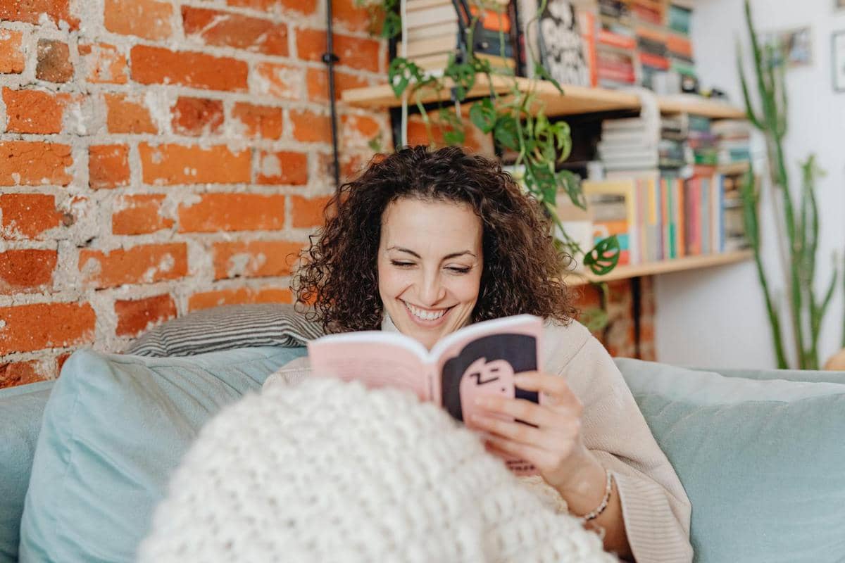 Seorang wanita tersenyum sambil membaca buku di sofa dengan latar dinding bata merah dan rak penuh buku di belakangnya.