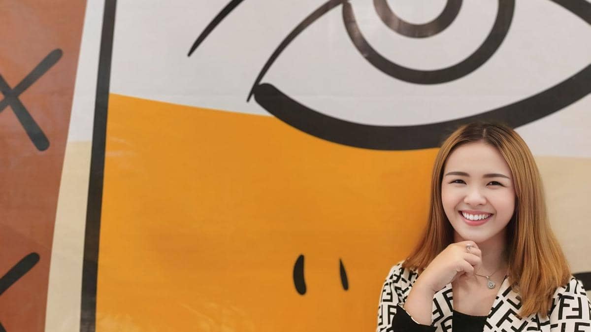 wanita berbaju hitam putih tersenyum dengan latar belakang mural berwarna oranye dan putih