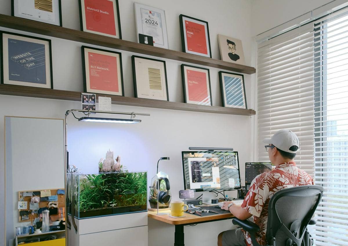Seseorang bekerja di meja dengan komputer ganda di ruangan modern yang memiliki akuarium tanaman air dan dinding berisi bingkai penghargaan.