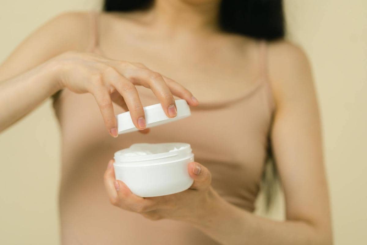 Seorang perempuan membuka wadah krim putih yang berisi pelembap atau soothing agent dengan latar warna netral.