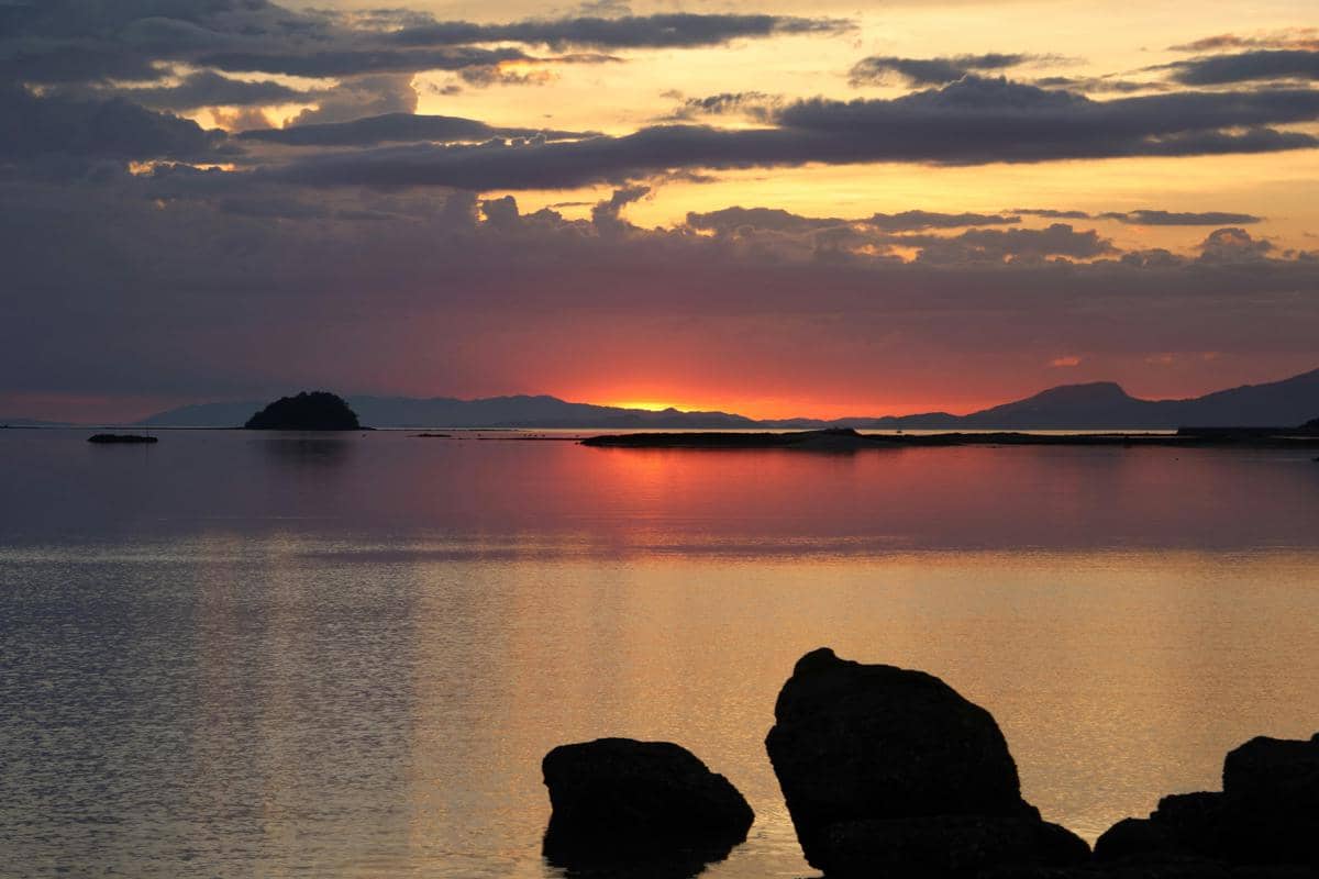 Ilustrasi Pantai Pulau Merah Banyuwangi dengan bukit kecil ikonik di tengah laut saat matahari terbenam.