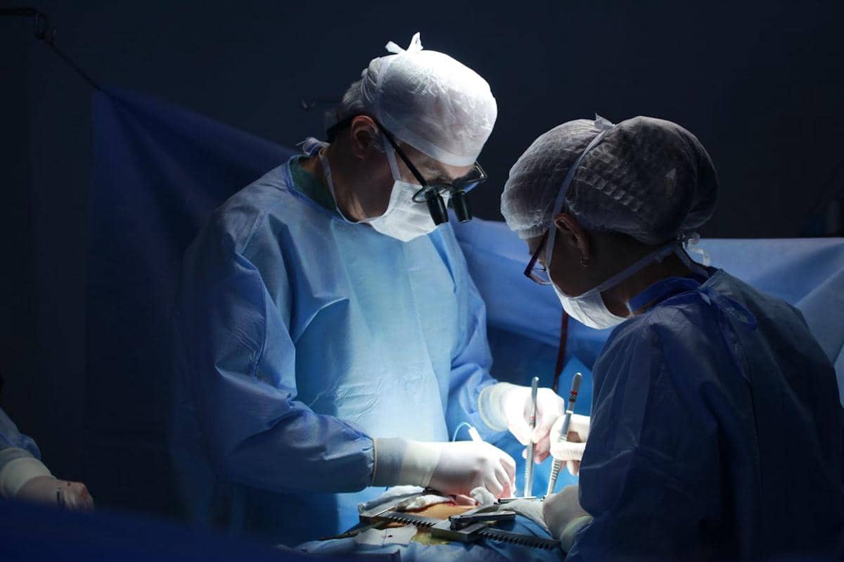 Tim dokter mengenakan pakaian bedah biru sedang melakukan operasi pada pasien di ruang operasi dengan pencahayaan fokus.