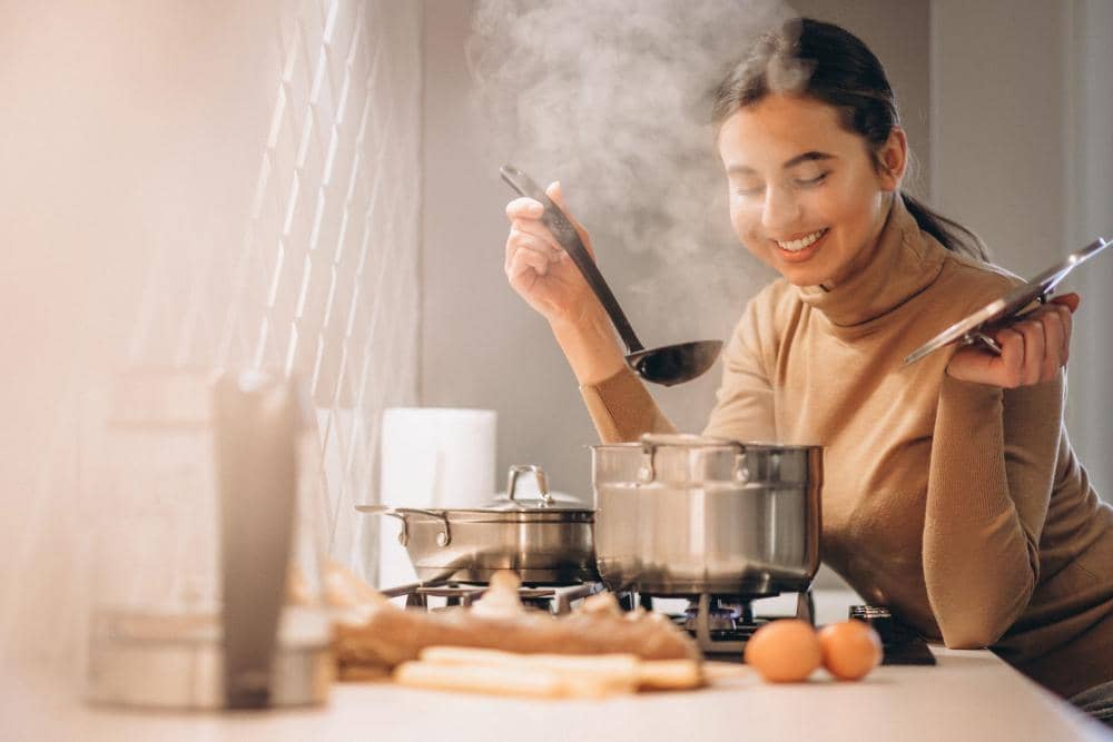 Seorang wanita tersenyum sambil mencicipi masakan dari panci di dapur modern dengan uap panas yang mengepul dari masakan.
