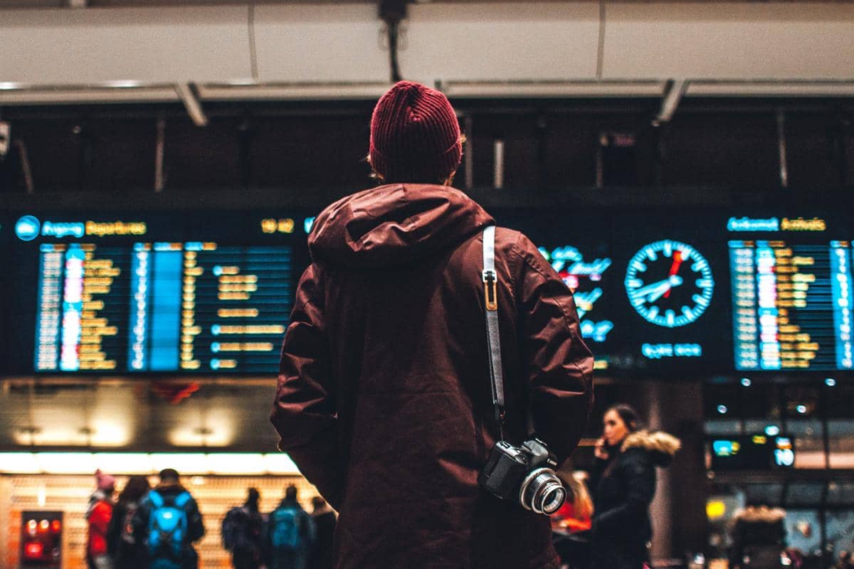 Seorang pria mengenakan jaket cokelat dan topi merah berdiri di depan papan jadwal keberangkatan di bandara dengan kamera tergantung di bahunya.