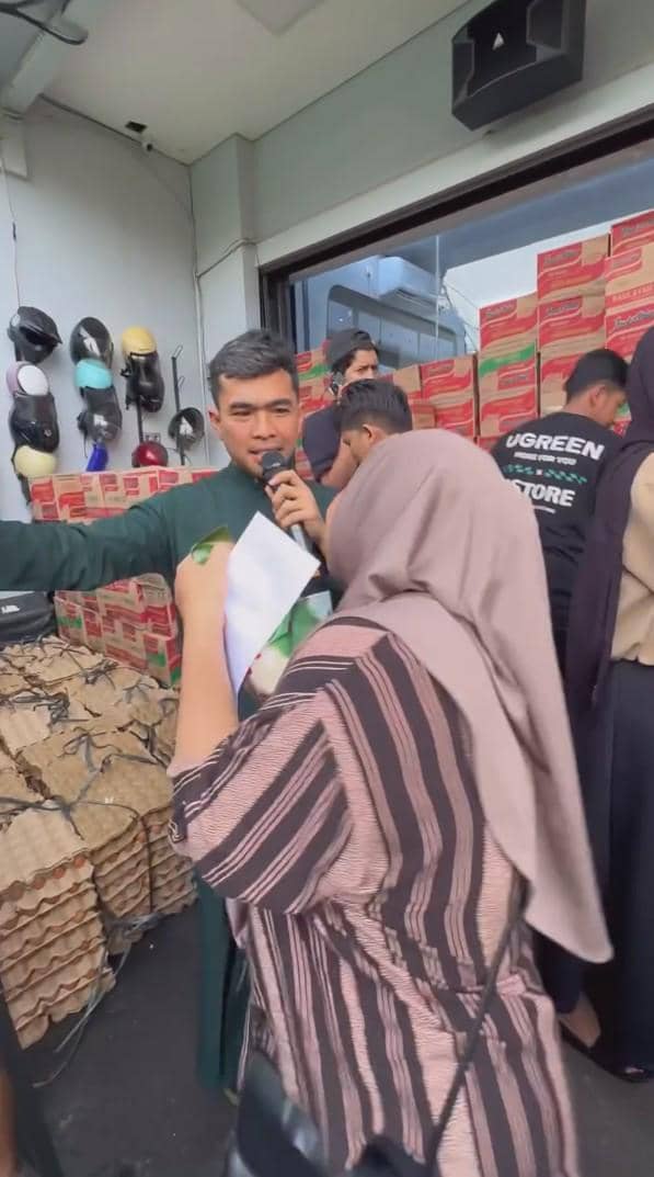 Putra Siregar sedang membagikan berbagai macam sembako, kardus mi instan dan telur terlihat menumpuk di belakang