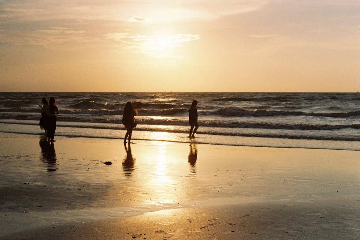 Beberapa orang berjalan di tepi pantai saat matahari terbenam dengan pantulan cahaya oranye di permukaan air dan pasir basah.