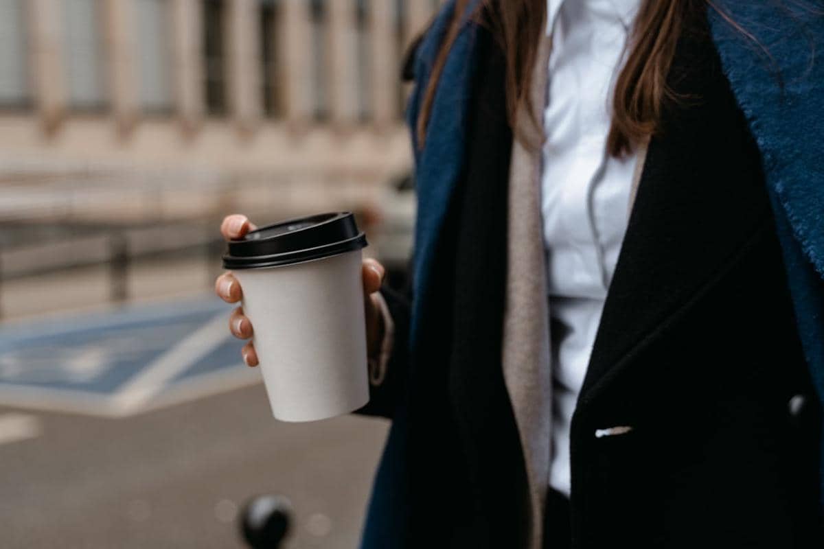 Seseorang mengenakan jas biru dan hitam memegang gelas kopi kertas dengan tutup hitam di luar ruangan.