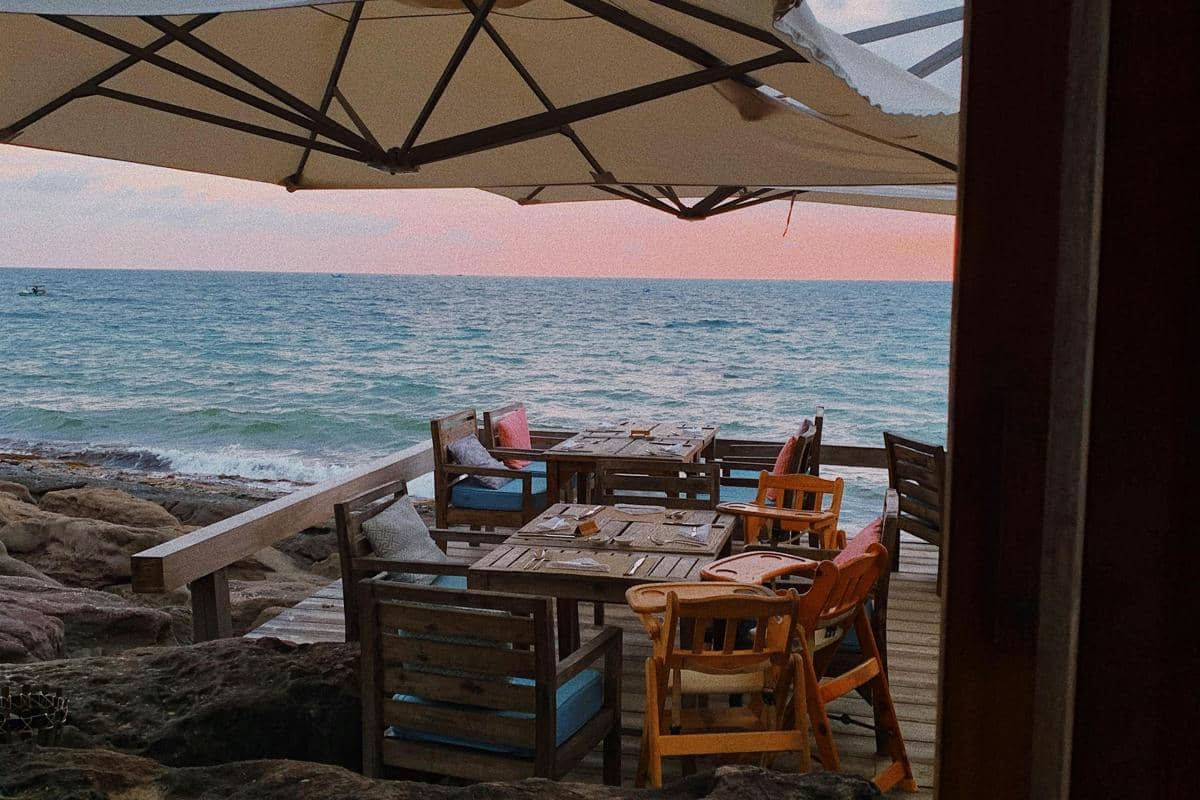Ilustrasi cafe tepi pantai dengan meja makan menghadap laut saat sunset.