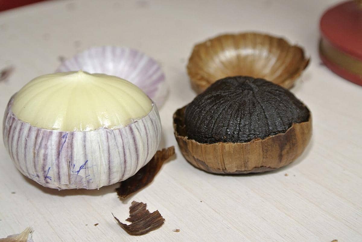 Beberapa umbi bawang hitam utuh dan siung bawang hitam kupas tersusun di atas latar putih yang bersih.