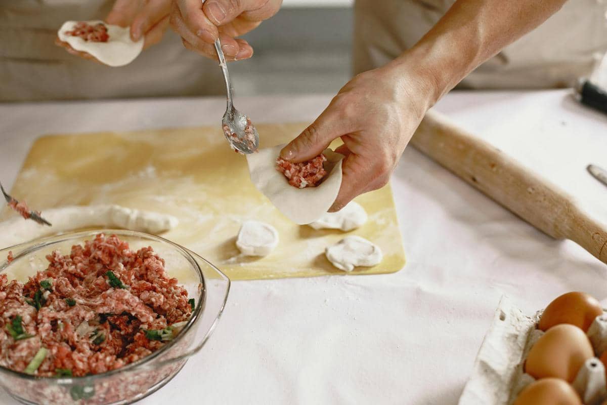 Tangan seseorang sedang mengisi kulit dimsum dengan adonan daging cincang di atas meja dapur yang dipenuhi bahan masakan.