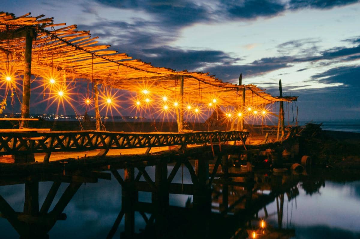 Ilustrasi warung seafood sederhana di tepi laut Banyuwangi dengan suasana senja.