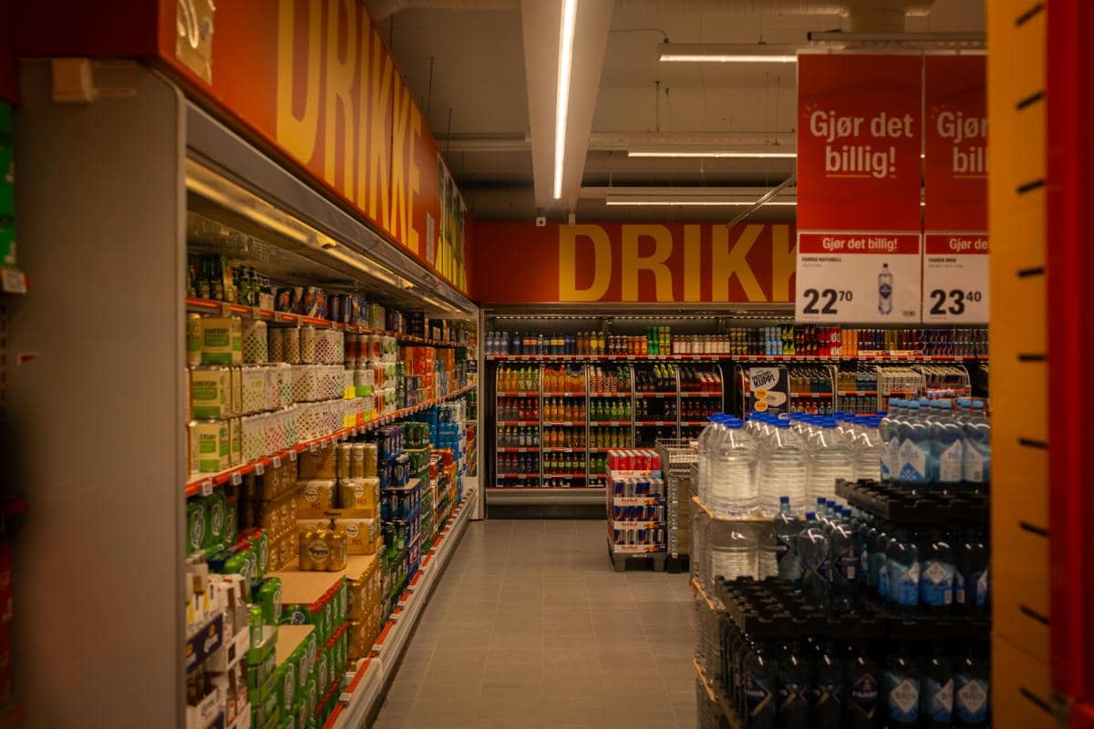 Deretan rak minuman dan air kemasan di dalam supermarket dengan pencahayaan hangat dan papan harga promosi berwarna merah.