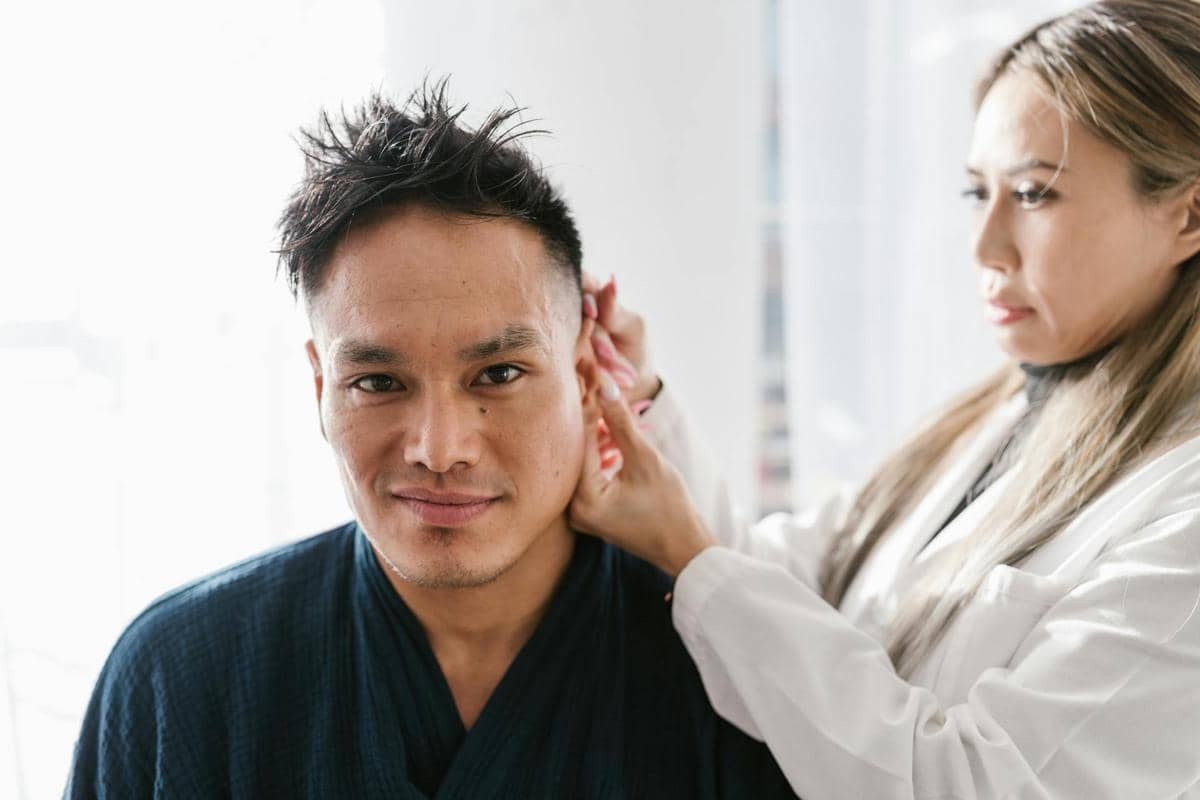 Seorang dokter memeriksa telinga seorang pria di ruang periksa dengan pencahayaan alami yang terang.