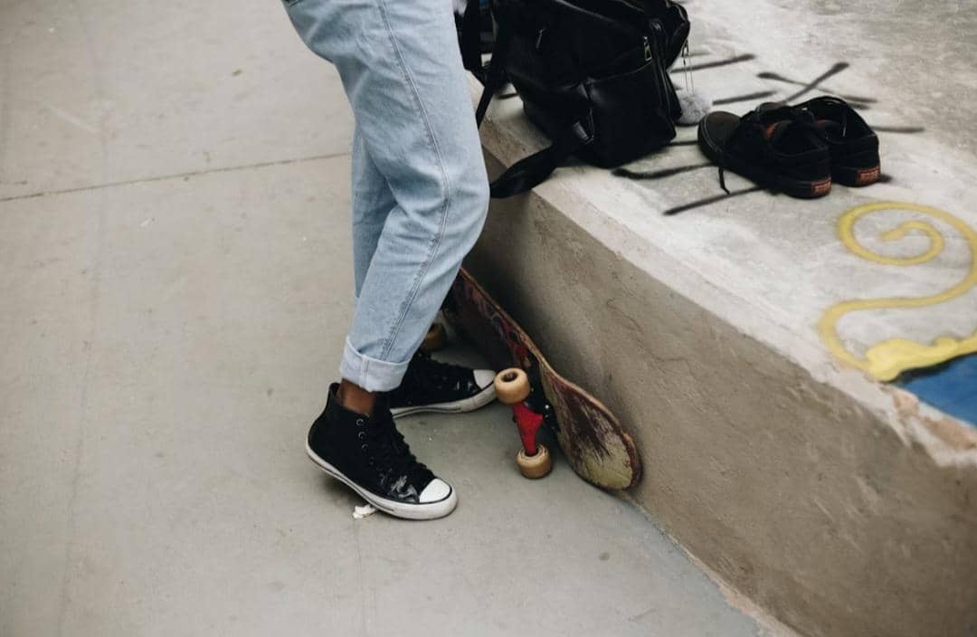 Seorang pria mengenakan celana jeans berwarna terang dan sepatu sneakers hitam berdiri di dekat skateboard dan tas hitam.