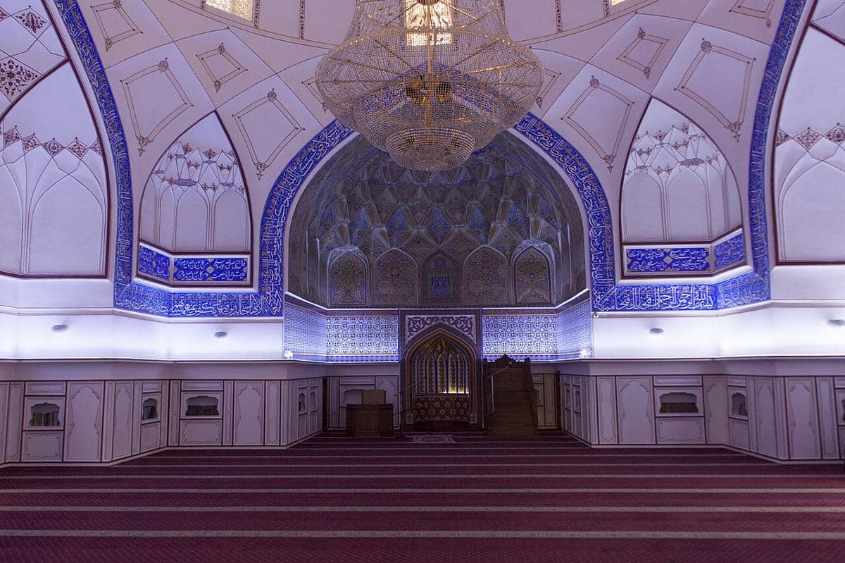 Interior aula salat Masjid Bolo Hauz dengan mihrab, mimbar kayu, dan lampu gantung di bagian tengah ruangan.