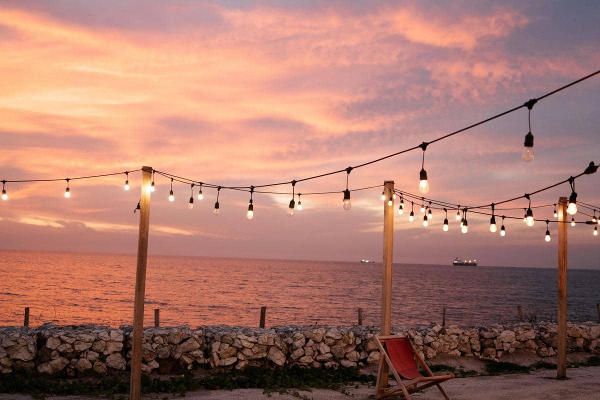 Ilustrasi suasana santai di Pantai Kenjeran Lama dengan latar Jembatan Suramadu saat sunset.