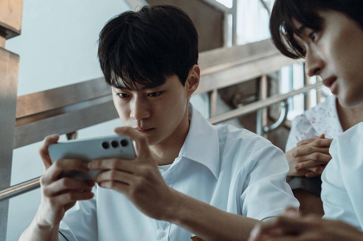 Seorang siswa berseragam putih menatap serius ke layar ponsel di tangannya, sementara temannya di sampingnya memperhatikan dengan cermat.