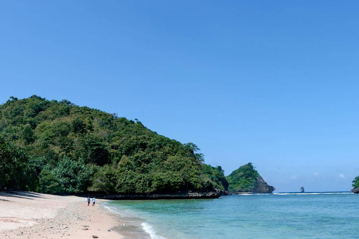 Ilustrasi Pantai Tiga Warna dengan air laut bergradasi biru–toska tampak jernih dengan suasana tenang alami.