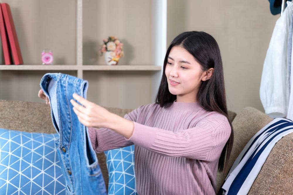 Seorang wanita duduk di sofa sambil memegang dan memperhatikan baju denim berwarna biru di ruang tamu yang rapi.