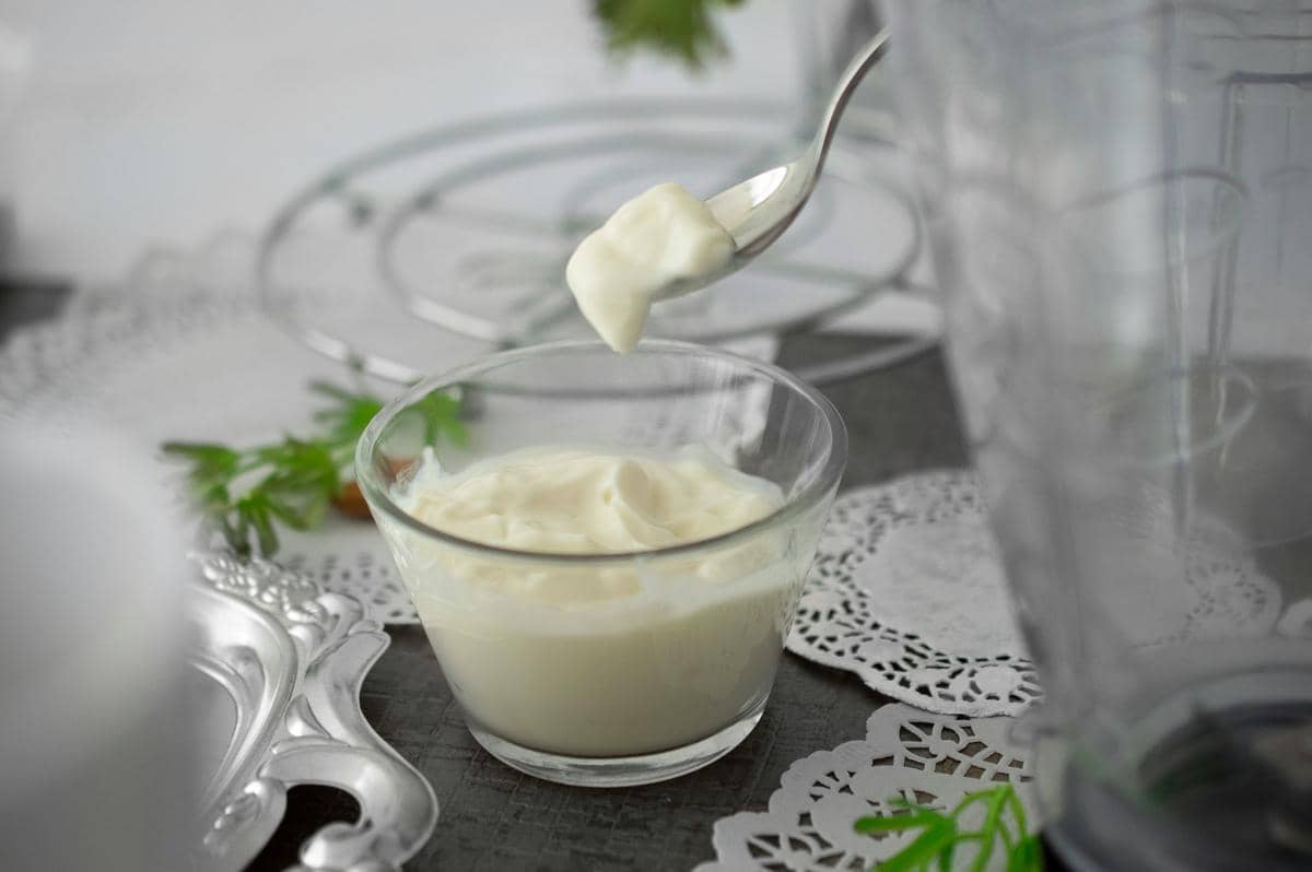 Semangkuk yoghurt putih kental di atas meja dengan sendok berisi yoghurt di atasnya, dikelilingi hiasan renda dan peralatan dapur.