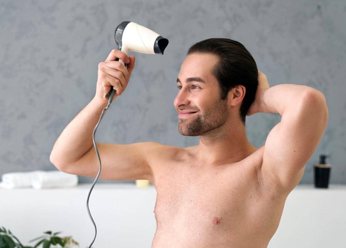 Seorang pria tanpa baju tersenyum sambil mengeringkan rambutnya menggunakan pengering rambut di kamar mandi modern.