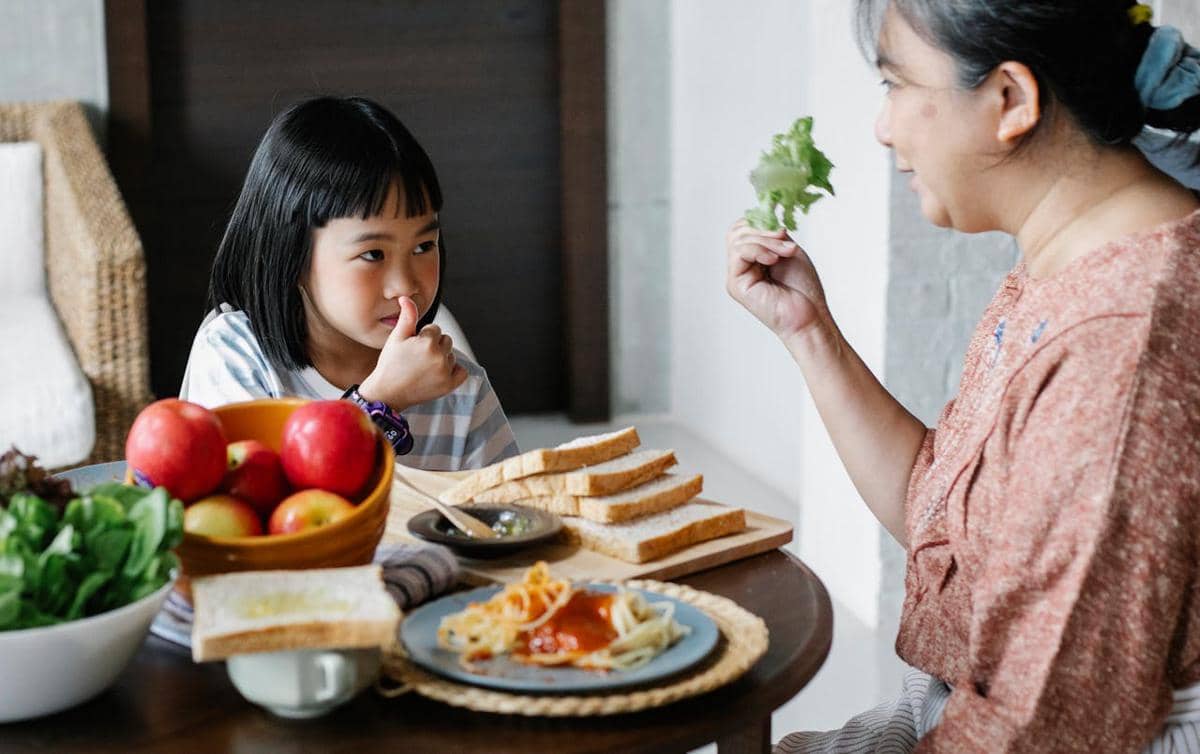 Seorang perempuan mengajak anak perempuan makan di meja makan dengan roti, pasta, dan buah-buahan di atas meja.