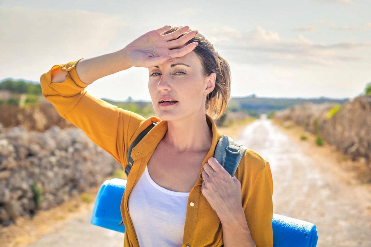 Seorang perempuan mengenakan kemeja kuning dan membawa ransel tampak kepanasan di jalan terbuka di bawah terik matahari.