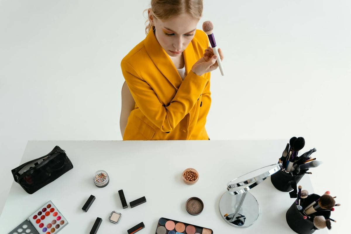Seorang wanita mengenakan blazer kuning sedang memegang kuas makeup di depan meja berisi berbagai perlengkapan kosmetik.