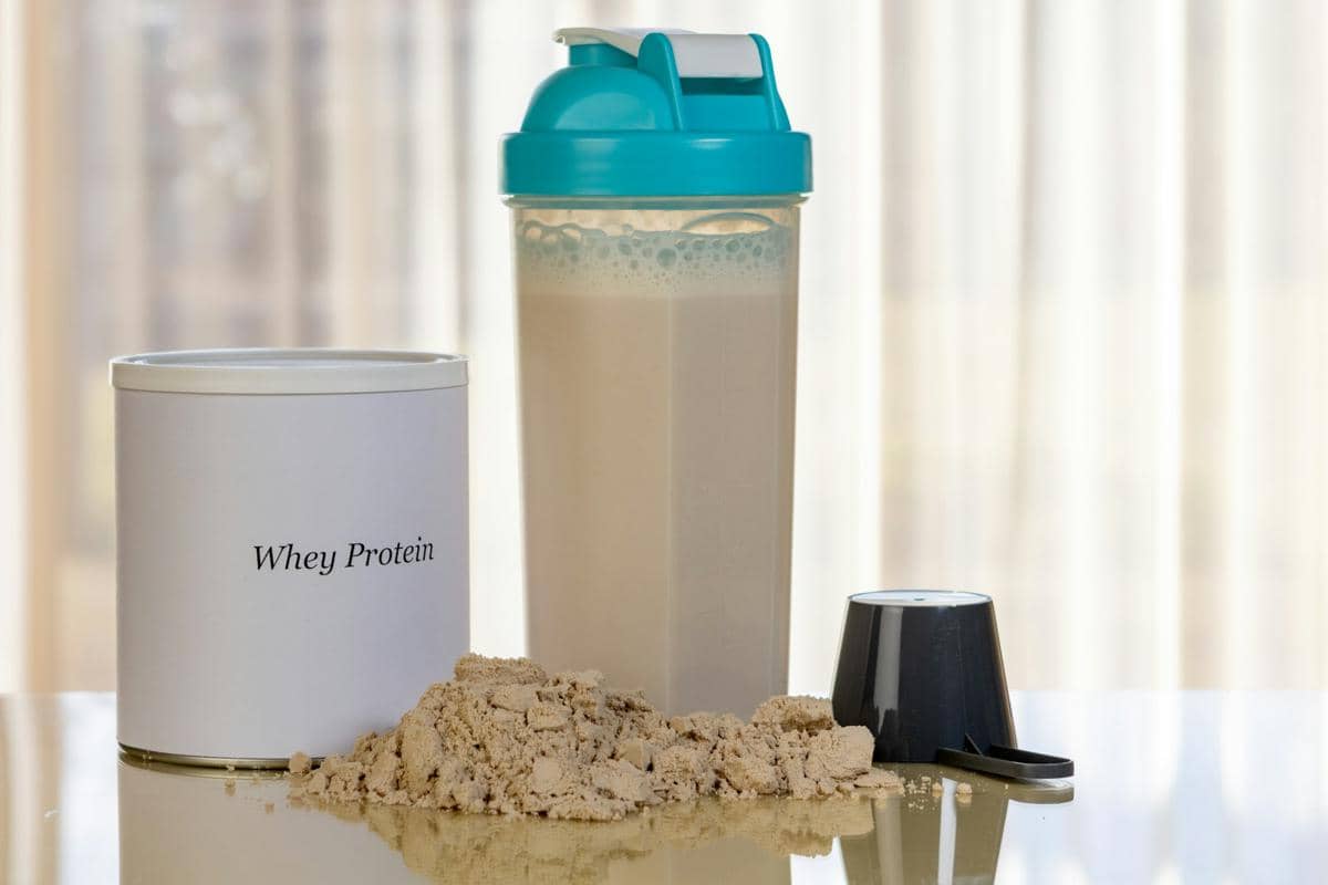 Bubuk whey protein berwarna cokelat muda di atas meja dengan wadah bertuliskan "whey protein" dan botol shaker berisi minuman protein.