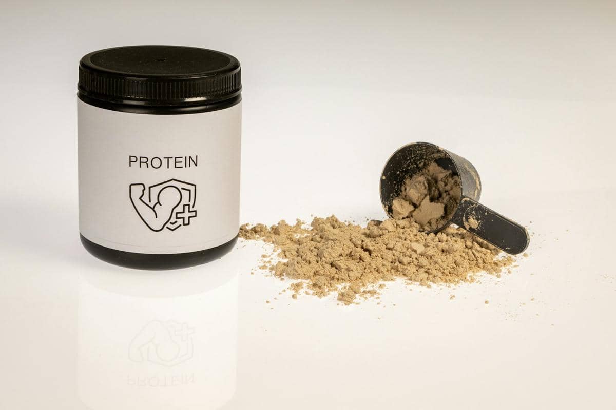 Wadah bubuk protein berlabel dengan sendok takar hitam berisi bubuk protein yang tumpah di permukaan putih.