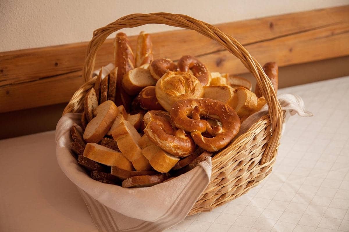 Keranjang anyaman berisi berbagai jenis roti seperti pretzel dan roti irisan yang tersusun rapi di atas kain putih.