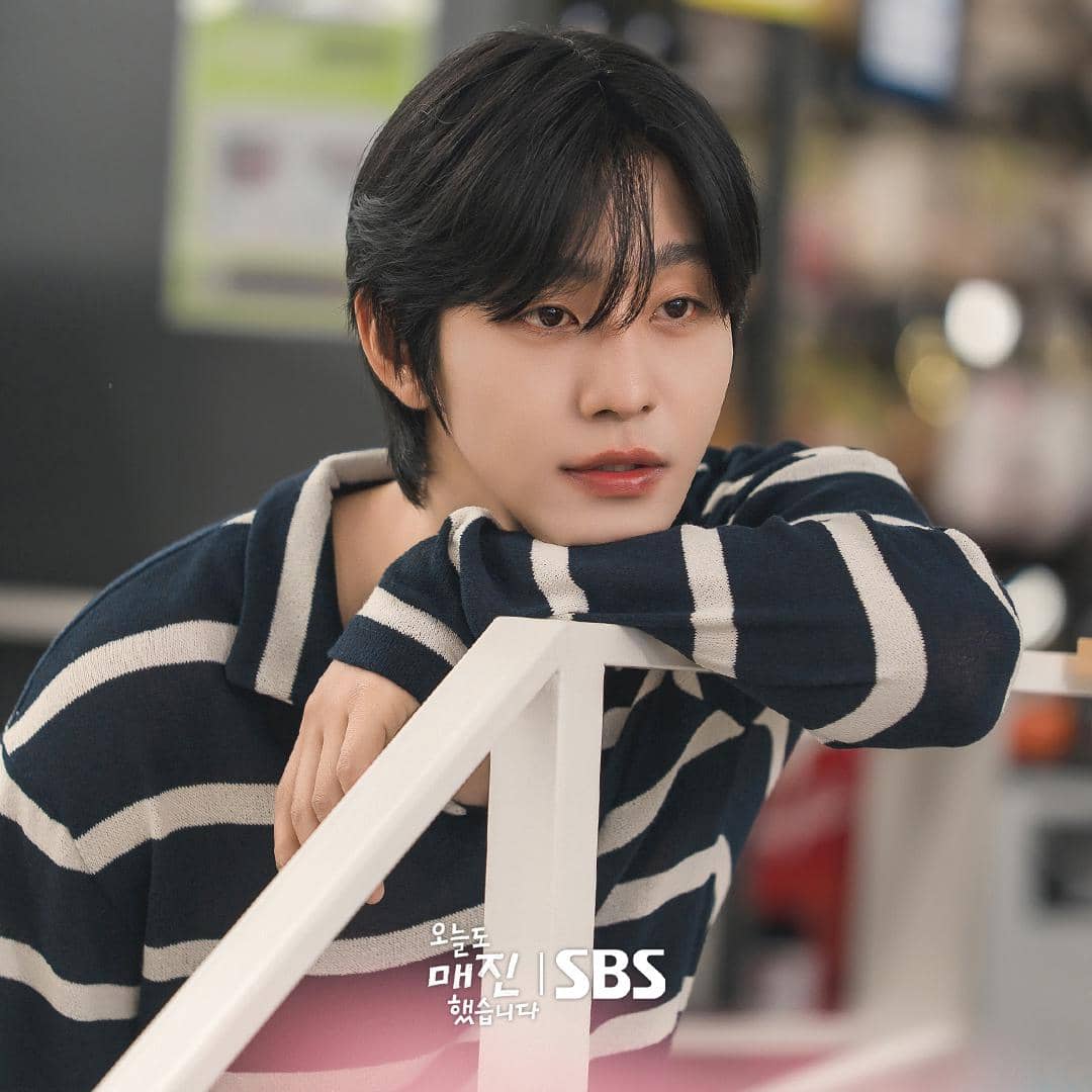 Seorang pria berambut hitam mengenakan sweter garis-garis biru dan putih bersandar di pagar dalam adegan drama Sold Out On You.