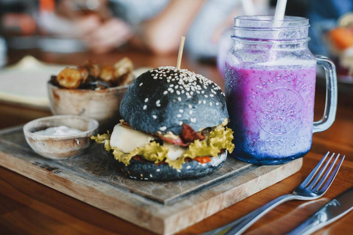 Burger dengan roti hitam berisi sayuran dan keju disajikan di papan kayu bersama minuman ungu dalam gelas kaca berpegangan.