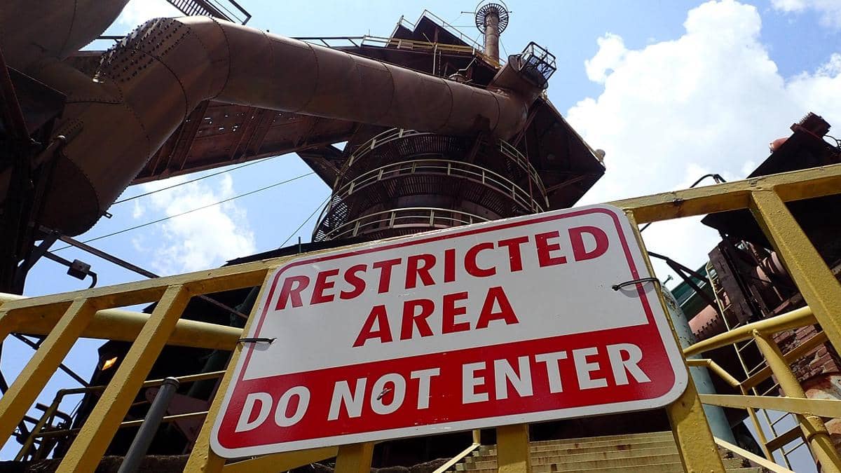 Papan peringatan bertuliskan 'Restricted Area Do Not Enter' di depan struktur industri logam besar dengan langit cerah di latar belakang.