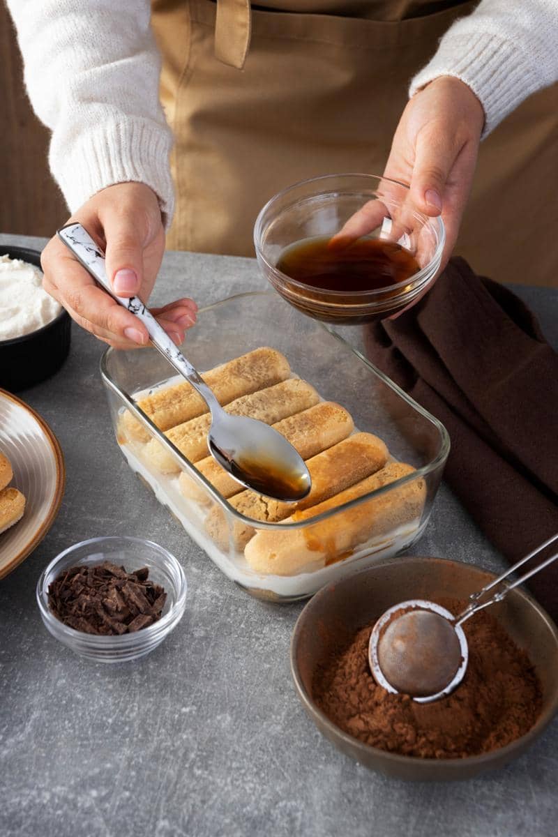 Tangan menuangkan kopi ke atas lapisan biskuit ladyfinger dalam wadah kaca untuk membuat tiramisu di atas meja dapur.