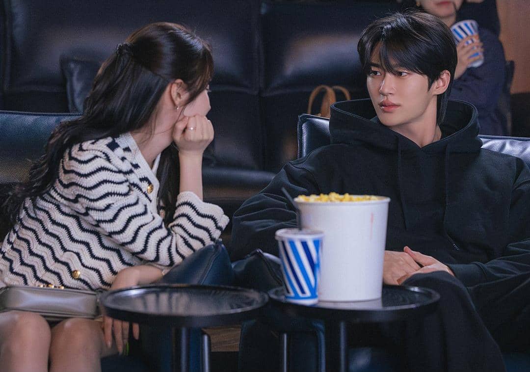 Seorang pria dan wanita duduk berdampingan di bioskop dengan popcorn dan minuman di meja kecil di antara mereka.