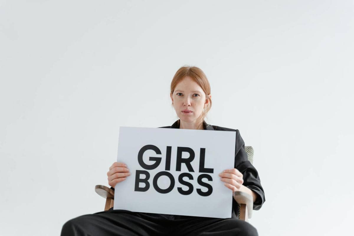 Seorang wanita berambut merah duduk di kursi sambil memegang papan bertuliskan 'GIRL BOSS' dengan latar belakang putih minimalis.
