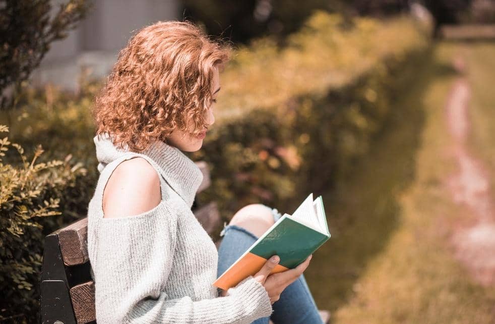 Seorang perempuan berambut keriting mengenakan sweter abu-abu duduk di bangku taman sambil membaca buku berwarna hijau.