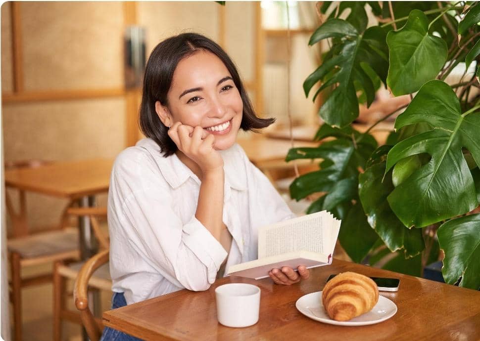 Seorang wanita tersenyum sambil membaca buku di kafe, dengan secangkir kopi dan croissant di atas meja dekat tanaman hijau.