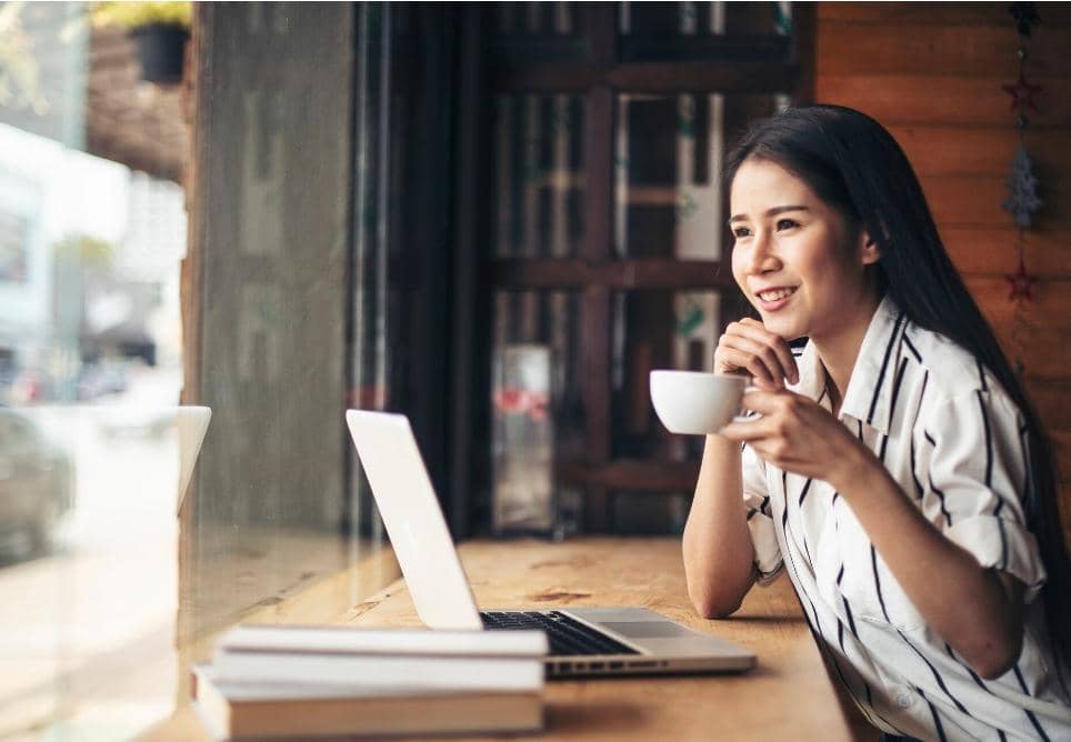 Seorang wanita duduk di kafe sambil memegang cangkir kopi dan menatap laptop di depannya dengan ekspresi bahagia.