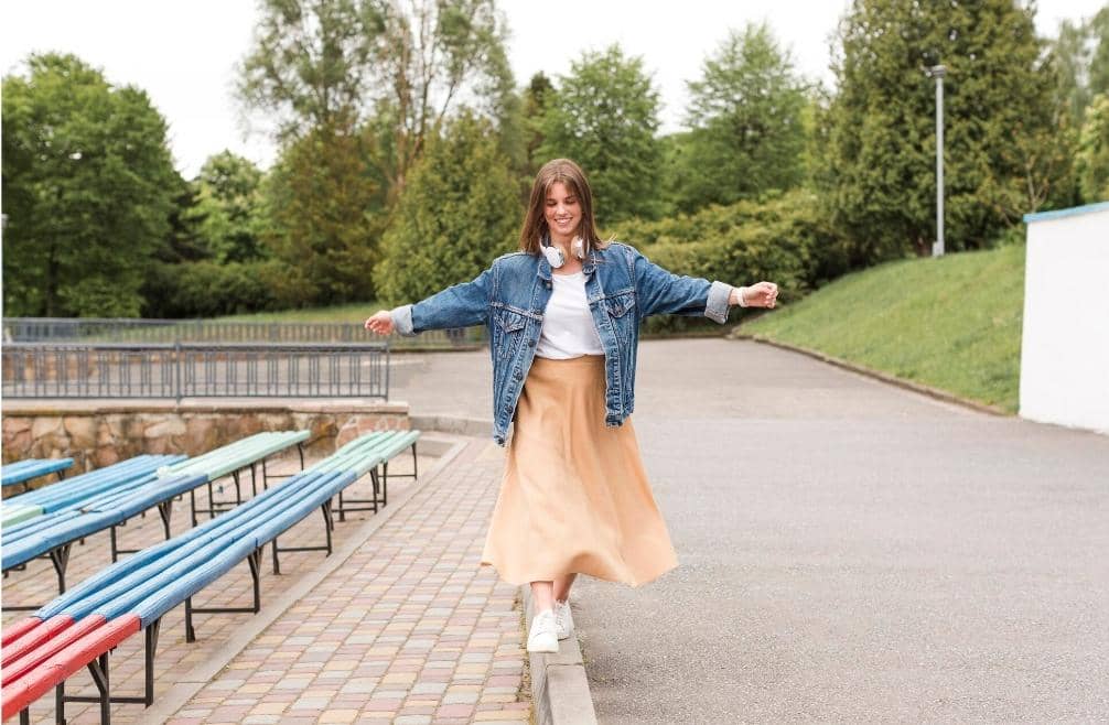 Seorang wanita muda mengenakan jaket denim dan rok panjang berwarna krem berjalan di tepi trotoar dengan latar taman hijau.