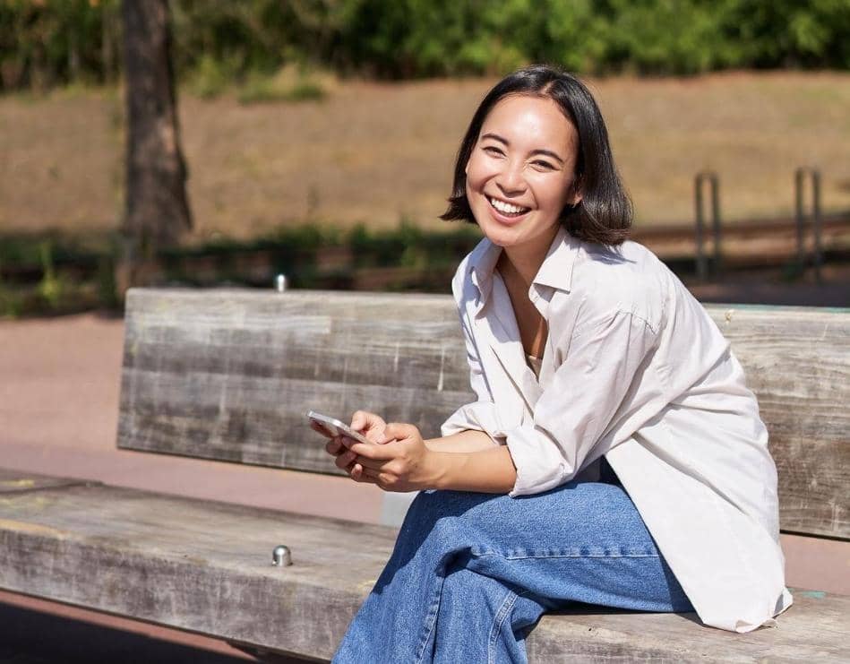 Seorang wanita muda tersenyum sambil duduk di bangku taman dan memegang ponsel, mengenakan kemeja putih dan celana jeans biru.