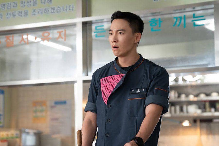 Seorang pria mengenakan seragam dapur berwarna gelap dengan detail merah muda berdiri di dapur, tampak terkejut dalam adegan drama Korea The Legend of Kitchen Soldier.