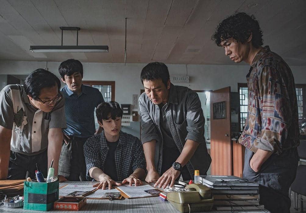 Lima pria sedang berdiskusi serius di sebuah ruangan kantor dengan dokumen dan alat tulis di atas meja, suasana tegang dan fokus.