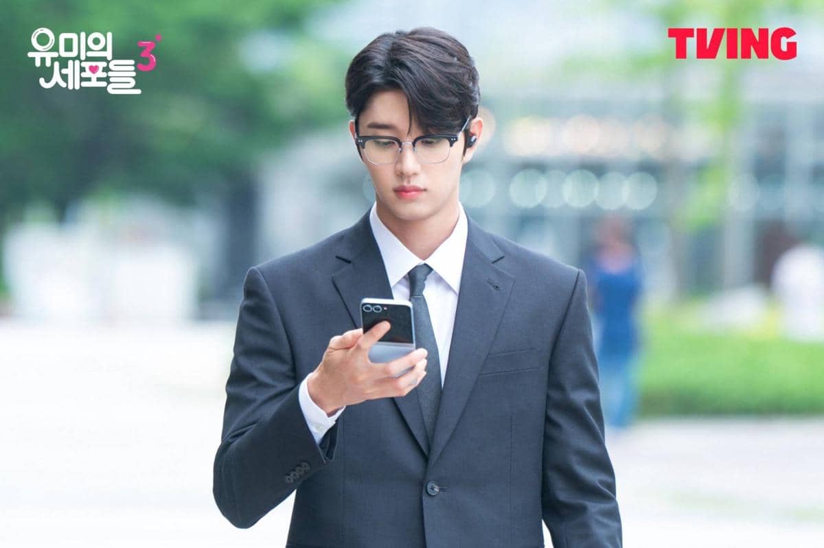 Seorang pria mengenakan setelan jas hitam dan kacamata sedang melihat ponsel di luar ruangan dalam cuplikan drama Korea Yumi's Cells 3.