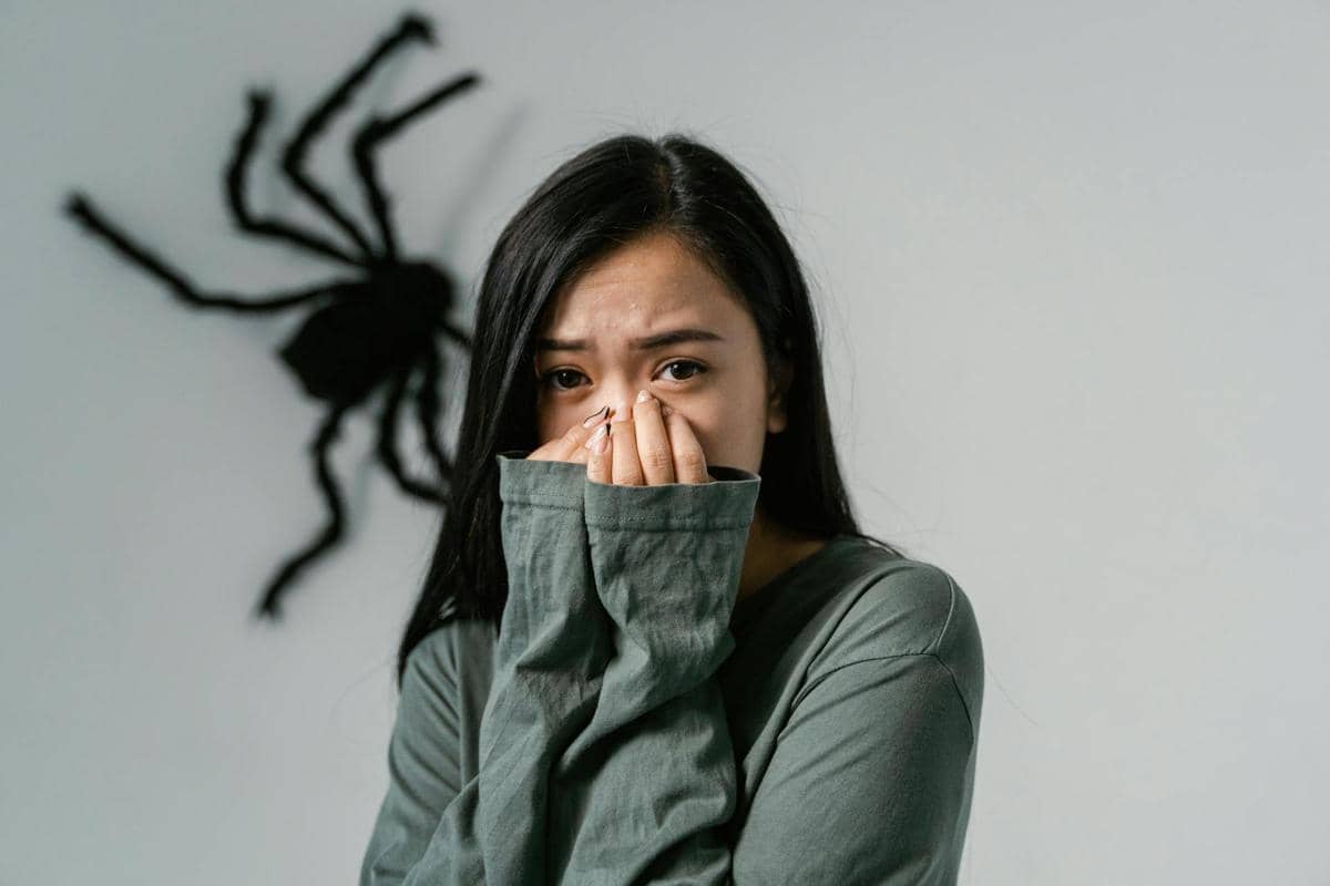 Seorang wanita berwajah cemas menutupi mulutnya dengan tangan, sementara di belakangnya tampak hiasan laba-laba besar di dinding.