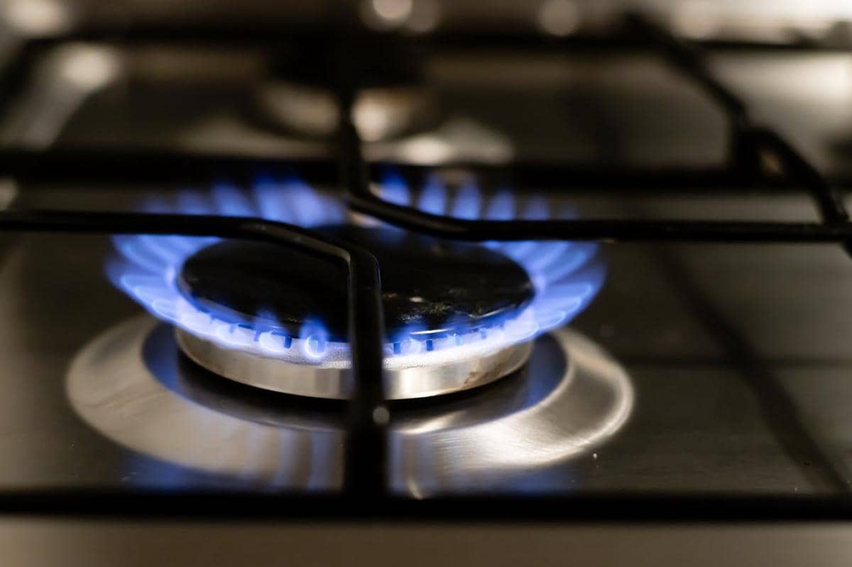 Api biru menyala di atas tungku kompor gas stainless steel dengan latar belakang dapur yang tampak bersih dan rapi.