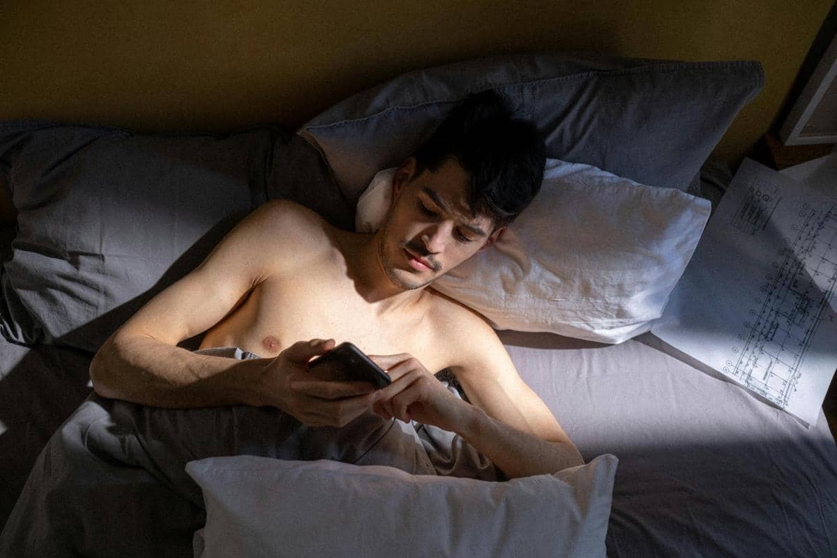 seorang pria berbaring di kasur sambil menatap layar ponsel di pagi hari
