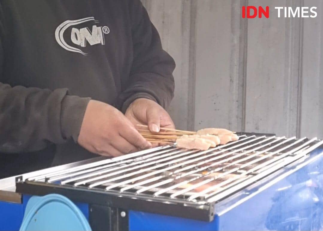 Seorang pedagang UMKM di Palembang memanggang sate ayam di atas panggangan logam biru untuk dijual kepada pelanggan.